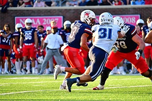 A football player in a blue jersey tackles an opponent in red during a game, with teammates and fans in the background.