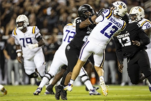 An LSU quarterback is tackled by a defensive player during a football game, with teammates in the background engaged in the play.