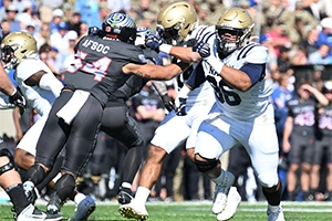 A football player in a white uniform blocks an opponent in black during an intense play on a sunny day, with a crowd visible in the background.