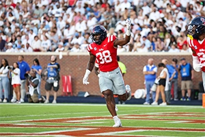 A college football player in a red jersey with the number 38 celebrates on the field, with a cheering crowd in the background.